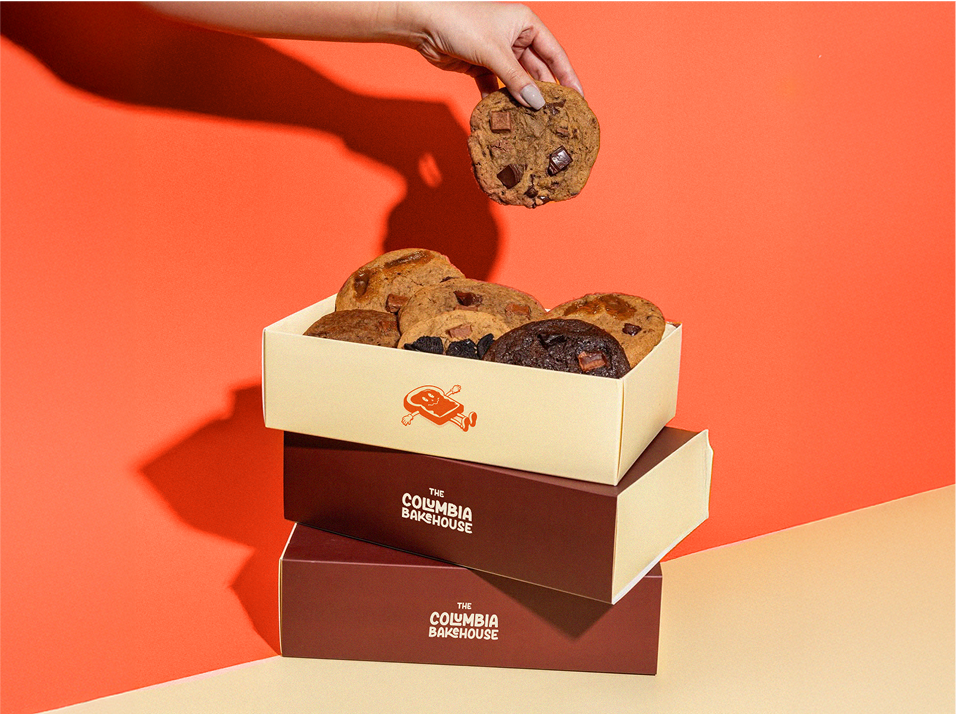 Three stacked Columbia Bakehouse cookie boxes with assorted cookies; hand reaching to grab a cookie